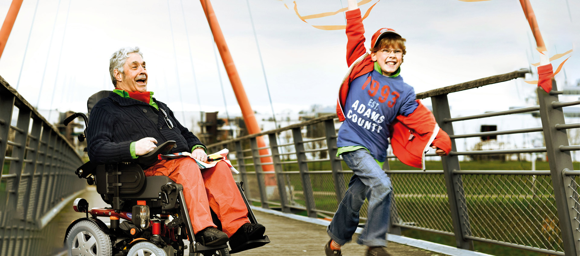 Man wearing orange pants, black jersey and orange and green scarf sits in a power wheelchair while a young boy in jeans and a blue and orange top runs and twirls an orange ribbon on a bridge