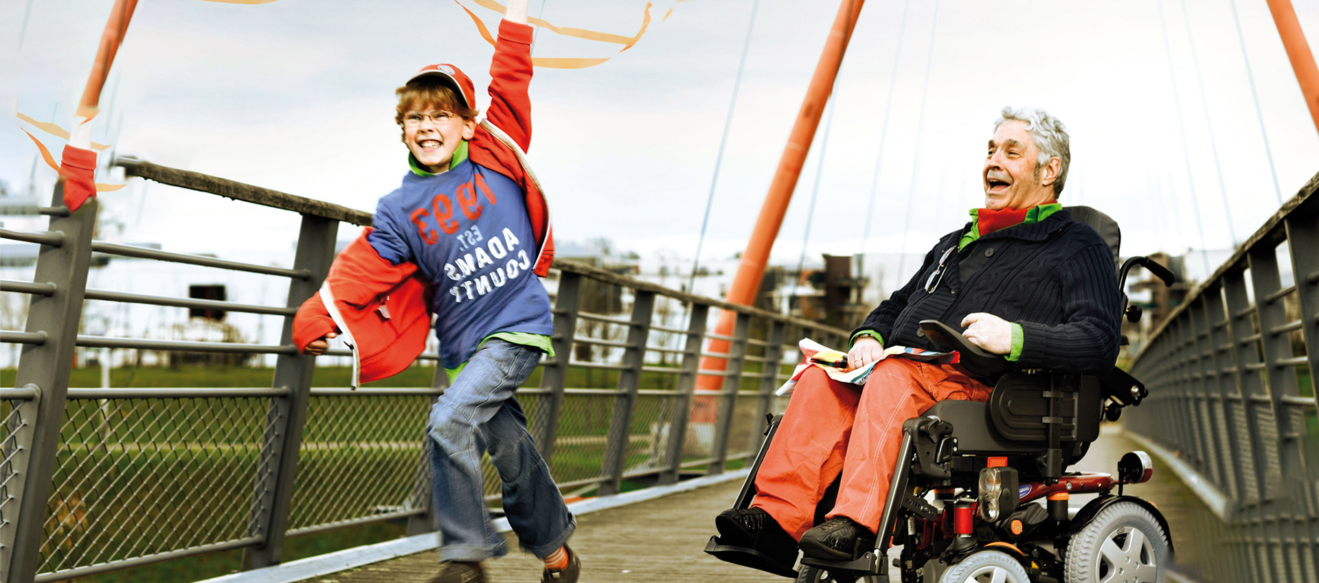 Man sits smiling in a power wheelchair while a boy runs and twirls an orange ribbon