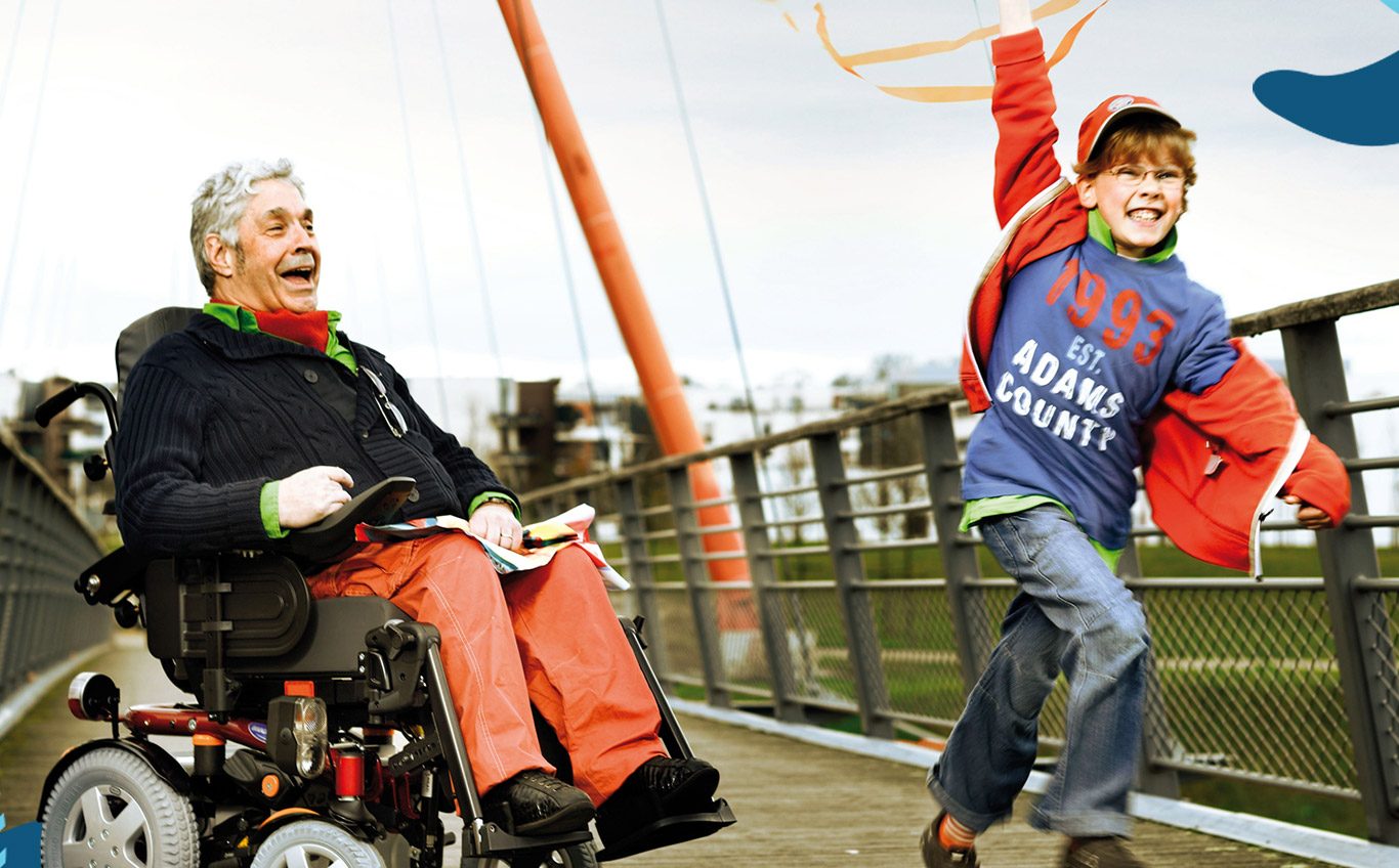 Man wearing orange pants, black jersey and orange and green scarf sits in a power wheelchair while a young boy in jeans and a blue and orange top runs and twirls an orange ribbon on a bridge