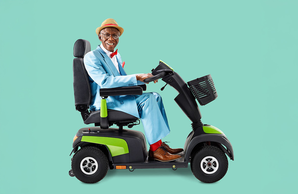 A man in a dapper blue suit and derby hat is smiling while he uses a MANTA powered mobility scooter