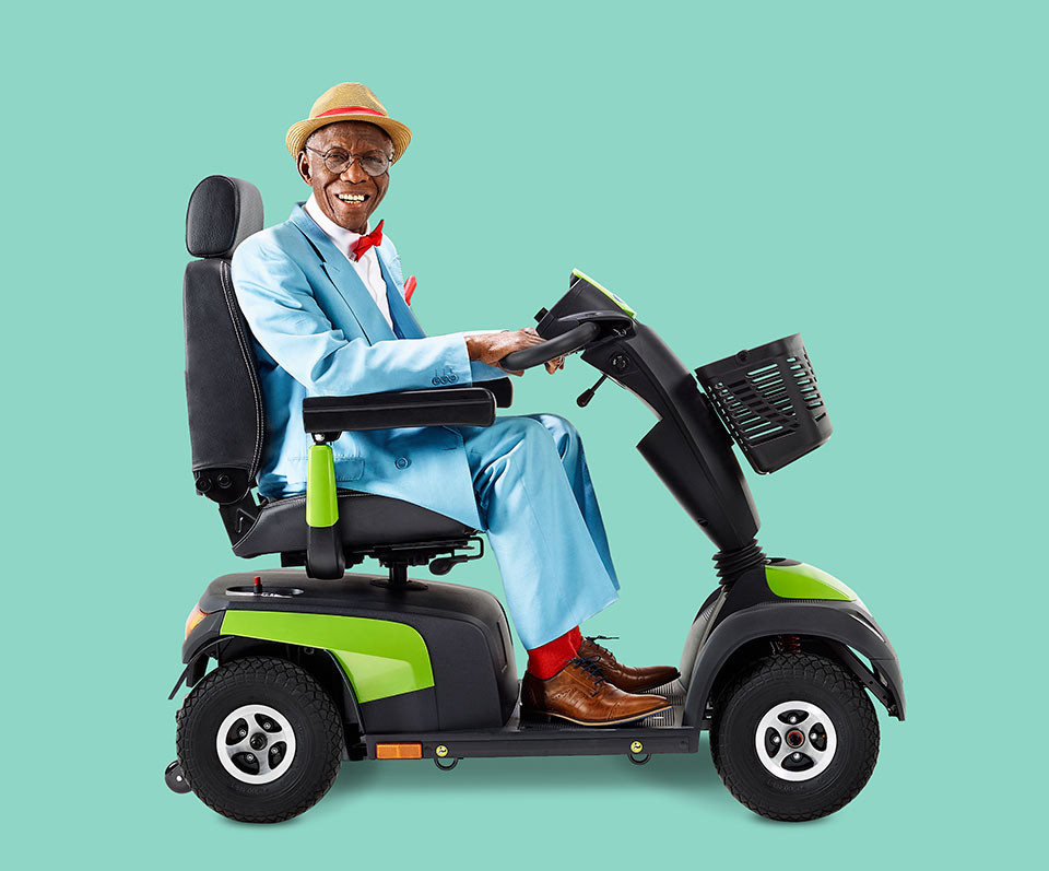 A man in a dapper blue suit and derby hat is smiling while he uses a MANTA powered mobility scooter