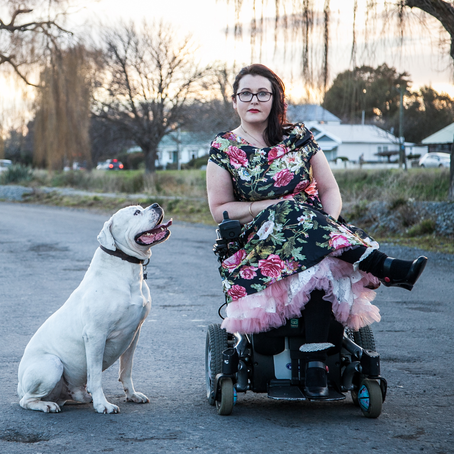 A woman wearing a floral dress with black boots on, sits in a powered wheelchair with white dog beside her