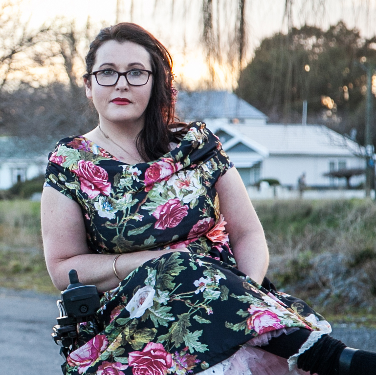 A woman wearing a rockabilly style dress with black boots on, sits in her Dynamic Controls powered wheelchair on a road while her white bull dog sits beside her